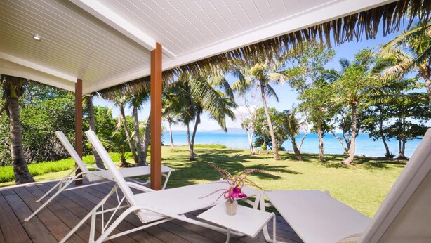 Ein überdachter Außenbereich mit weißen Lounge-Stühlen und einem Tisch, mit Blick auf eine tropische Landschaft mit Palmen und Meer.