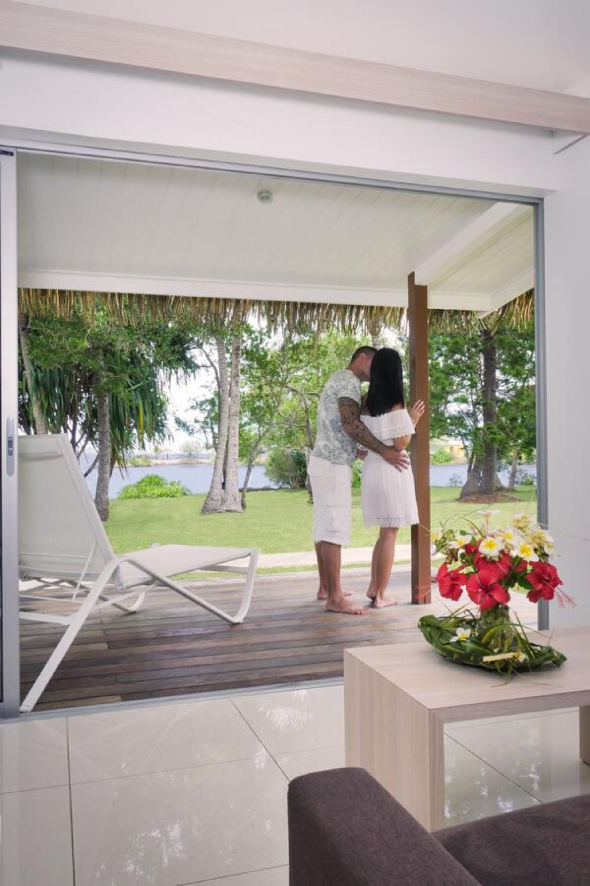 A couple embracing in a modern room with a glass door leading to a tropical garden and a view of the sea.