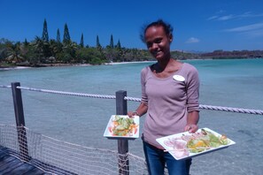 A person holding two plates of food on a rope bridge over a body of water with a clear sky and trees in the background. - Malabou Beach Hotel (Poum)
