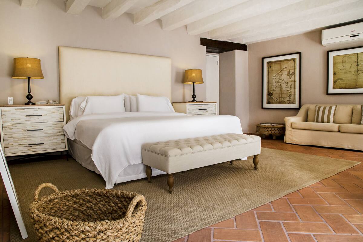 A bedroom with a large bed, two bedside tables with lamps, a bench, a woven basket, a sofa, and two framed maps on the wall.