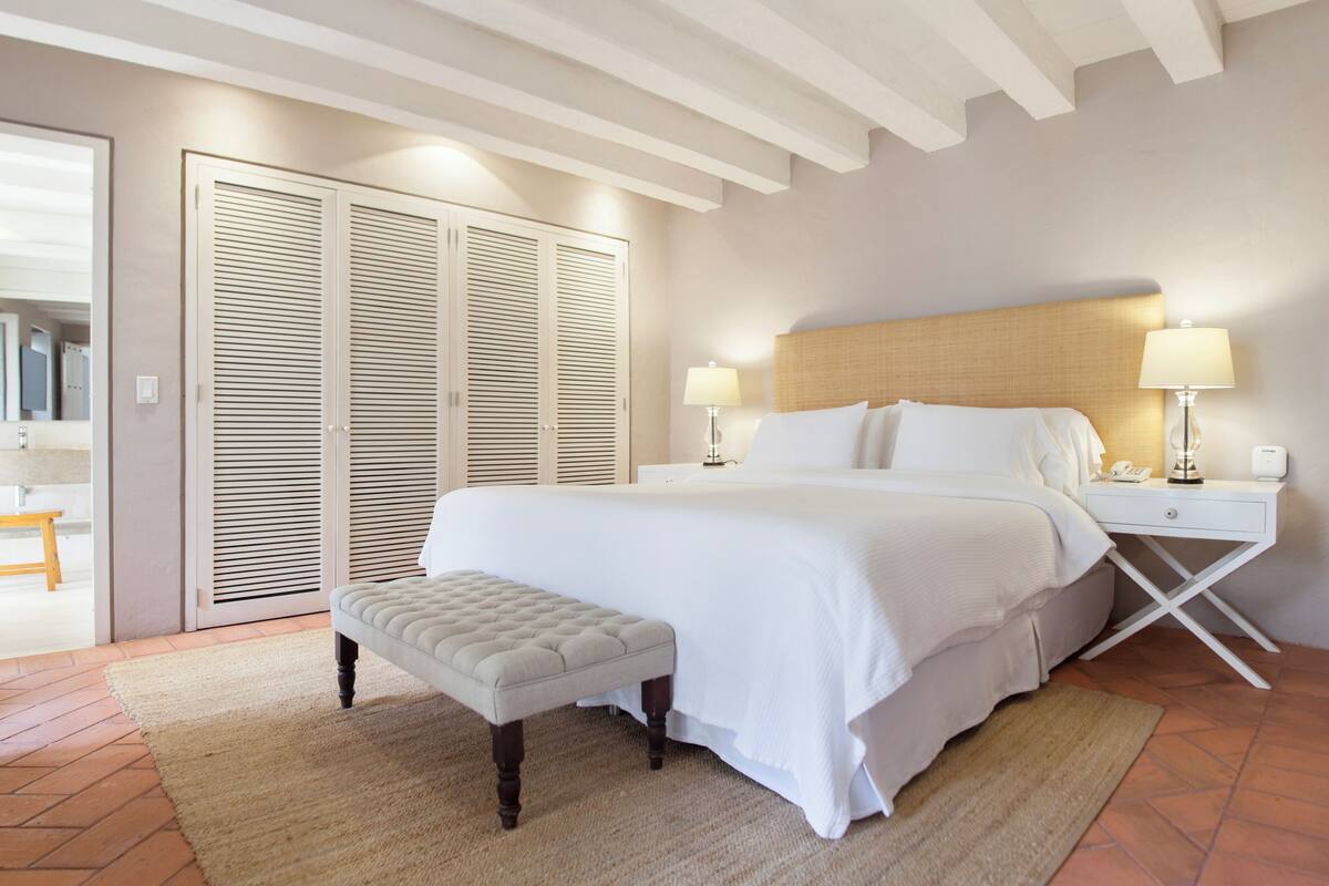 A hotel room with a large bed, two bedside tables with lamps, a wooden headboard, a bench, and a sliding closet.