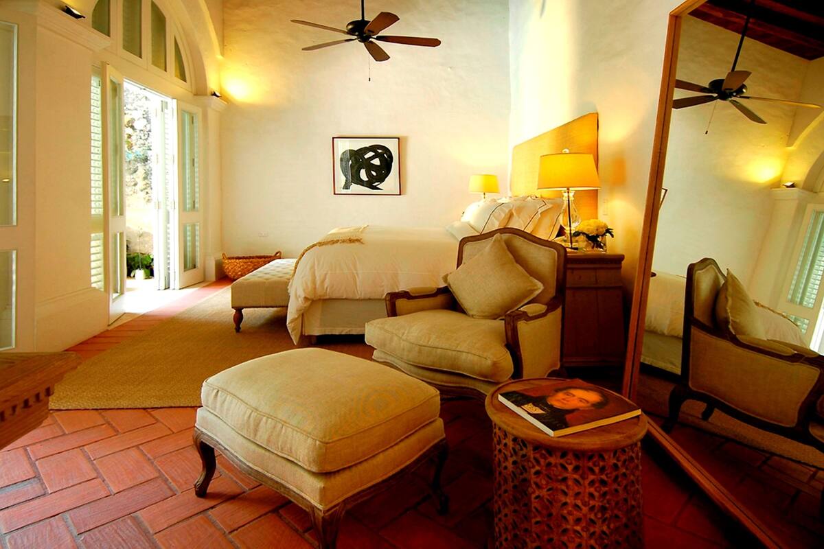 A hotel room with a bed, two armchairs, a small table with a book, a mirror, a ceiling fan, and a window with shutters.