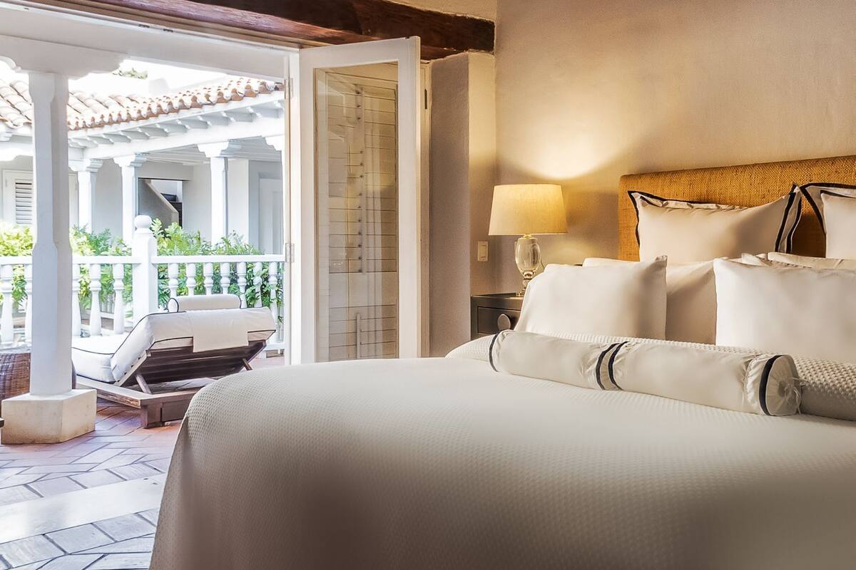 A hotel room with a large bed, a nightstand with a lamp, and a view of an outdoor patio with a chair and plants.