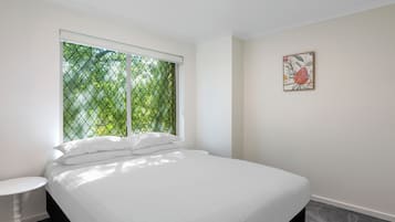 A bedroom with a bed, a round table, a window with a view of greenery, and a framed picture on the wall.