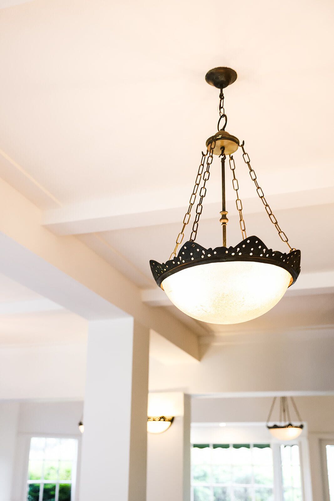 Un lustre doté d’une armature en métal noir et d’un abat-jour blanc, suspendu à un plafond dont les poutres sont apparentes.