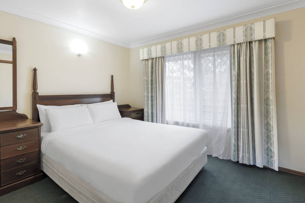 A neatly made bed with a wooden headboard, a nightstand with a lamp, and a window with patterned curtains.