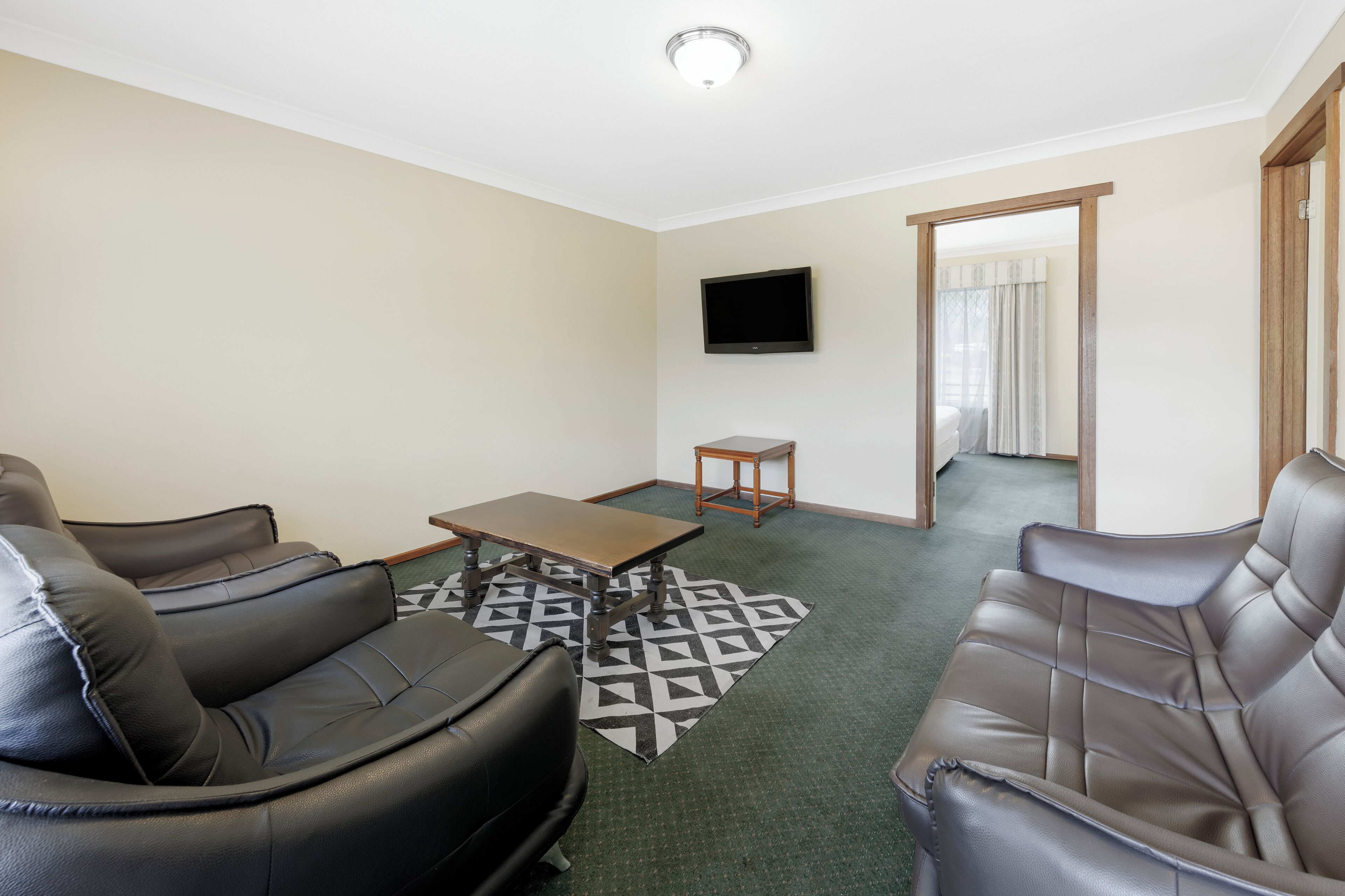 A living room with two black leather sofas, a wooden coffee table, and a patterned rug. A flat-screen TV is mounted on the wall, and a wooden door leads to another room.