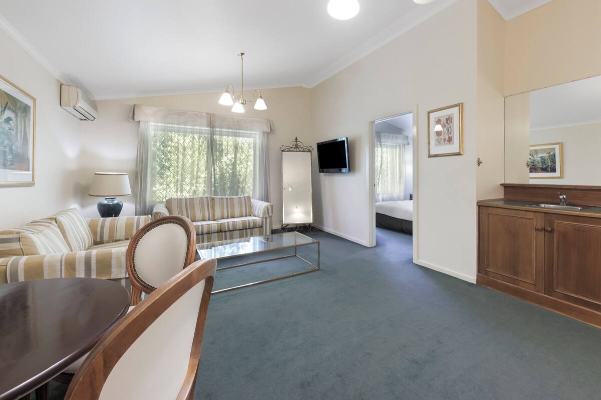 A living room with a sofa, a coffee table, and a TV. There is a kitchen area with wooden cabinets and a sink.