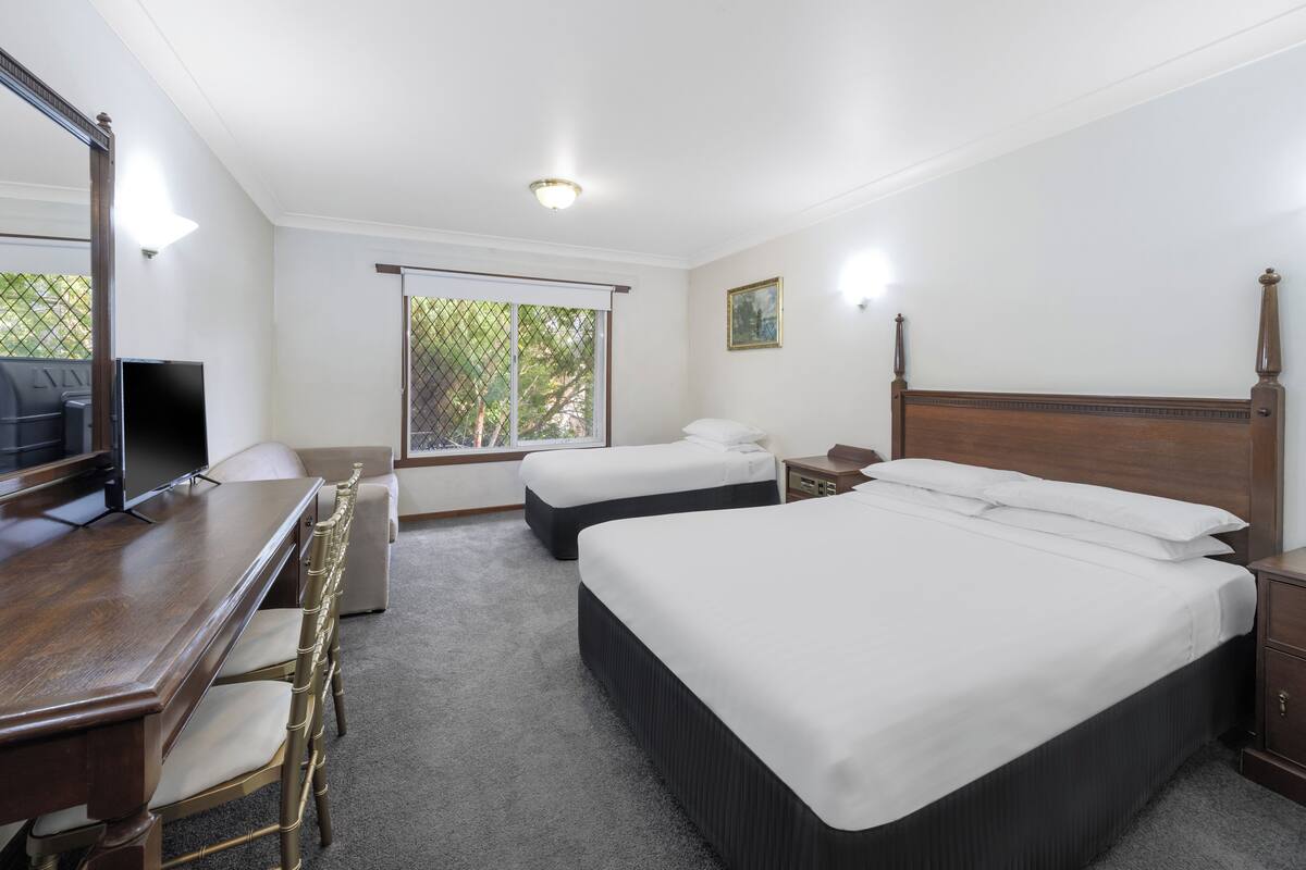 A hotel room with a wooden desk, two beds, a TV, and a window with a view of trees.