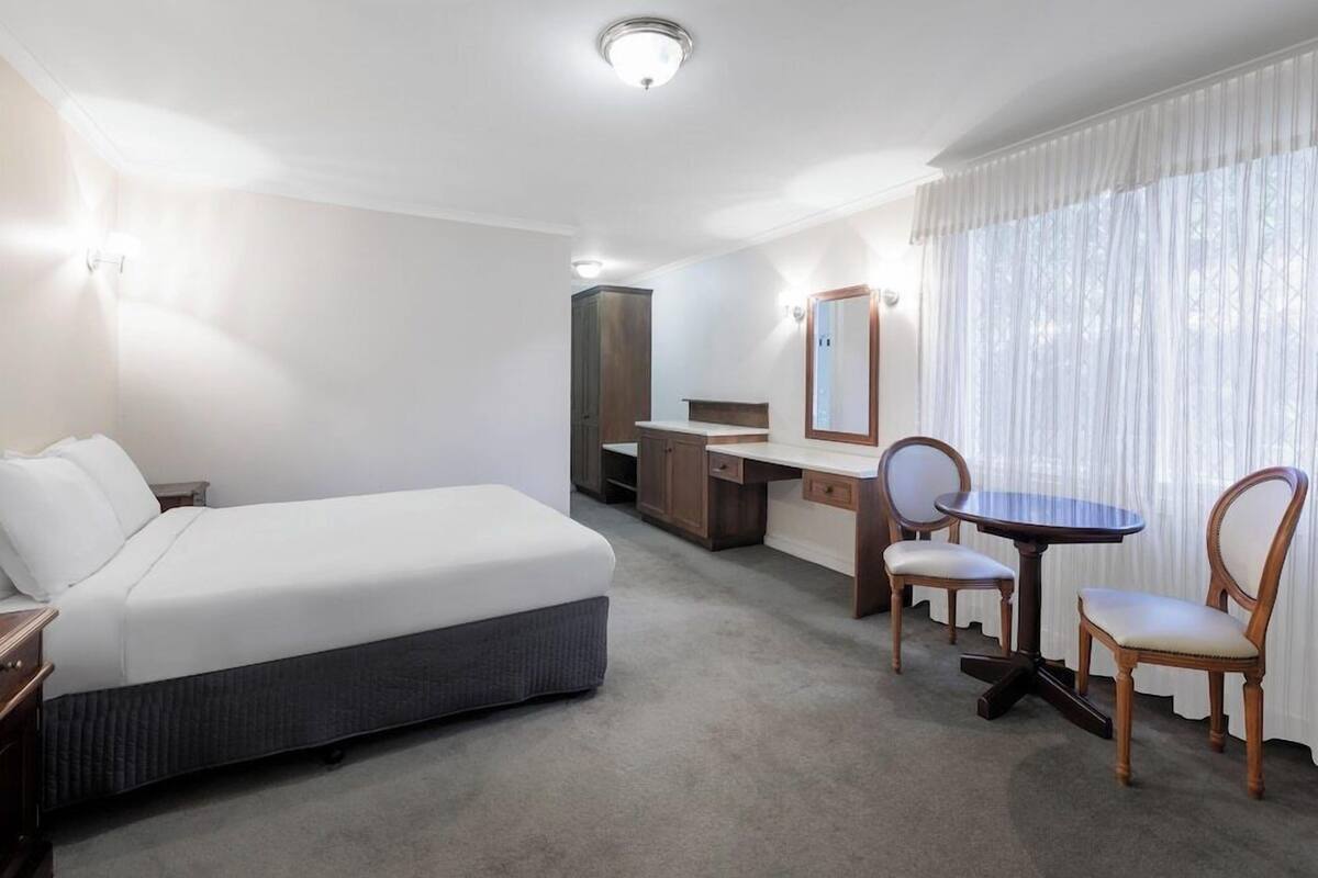 A hotel room with a bed, a desk, a chair, a table, and a large window with curtains.