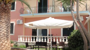 A patio with outdoor seating, a palm tree, and a building with a balcony. - Argo Hotel (Corfu)