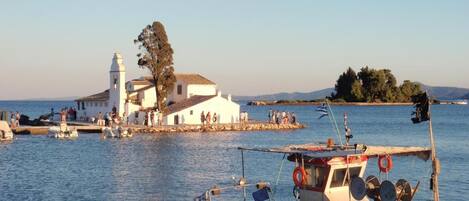 A coastal scene with a white building with a bell tower, a dock with boats, and a small island in the background.