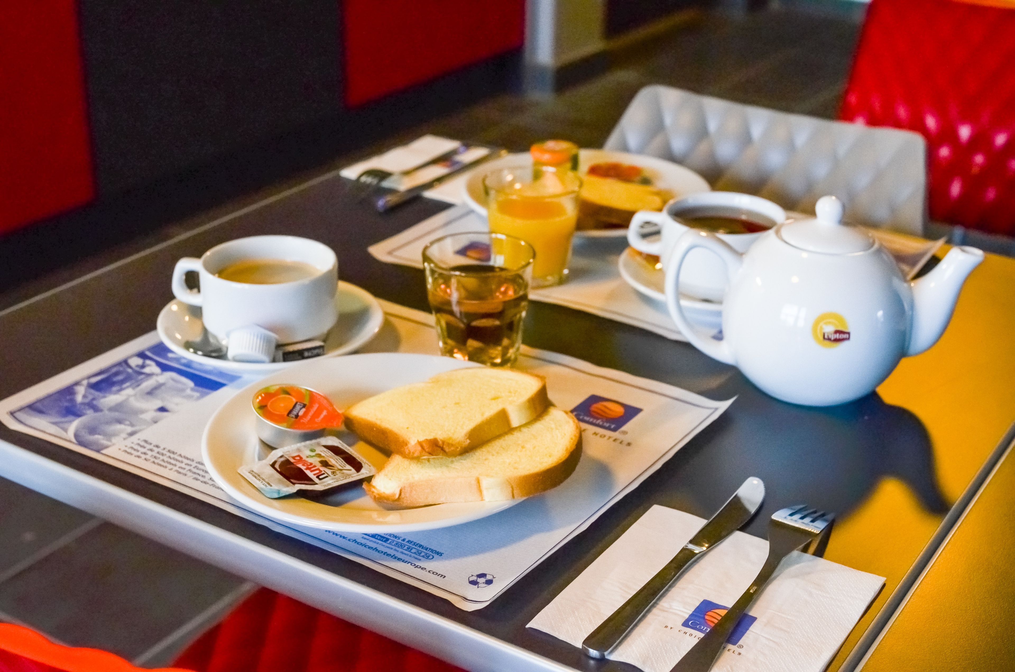 Ein Frühstückstisch mit einer Kaffeetasse, einem Orangensaftglas, einem Teekännchen, einer Toastscheibe mit Marmelade und einer kleinen Schale mit Obst.