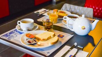 Ein Frühstückstisch mit einer Kaffeetasse, einem Orangensaftglas, einem Teekännchen, einer Toastscheibe mit Marmelade und einer kleinen Schale mit Obst.