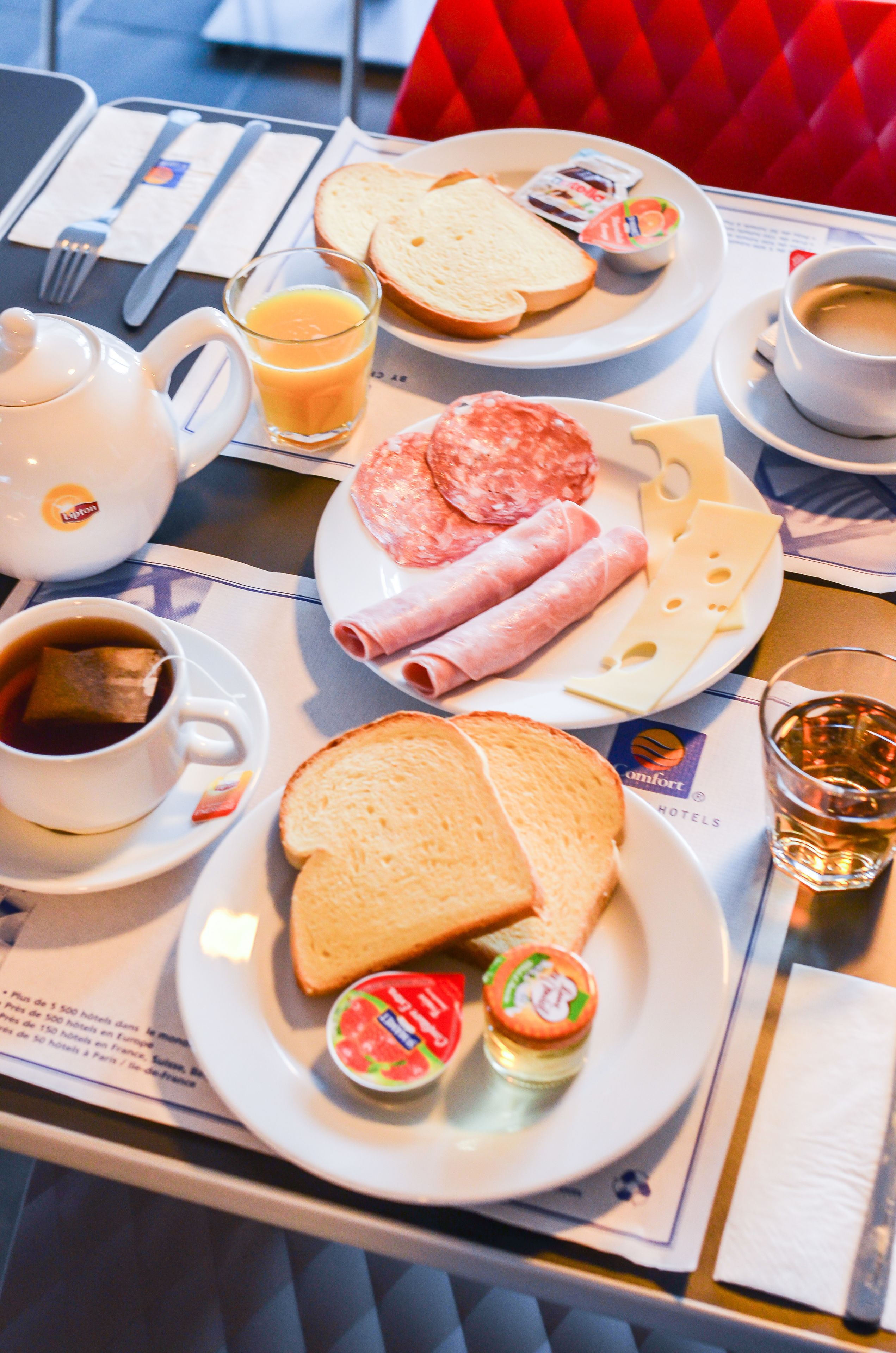 Un desayuno con un plato de pan en rodajas, queso, jamón y un recipiente pequeño de mermelada. También hay una taza de café y un vaso de zumo de naranja sobre la mesa.