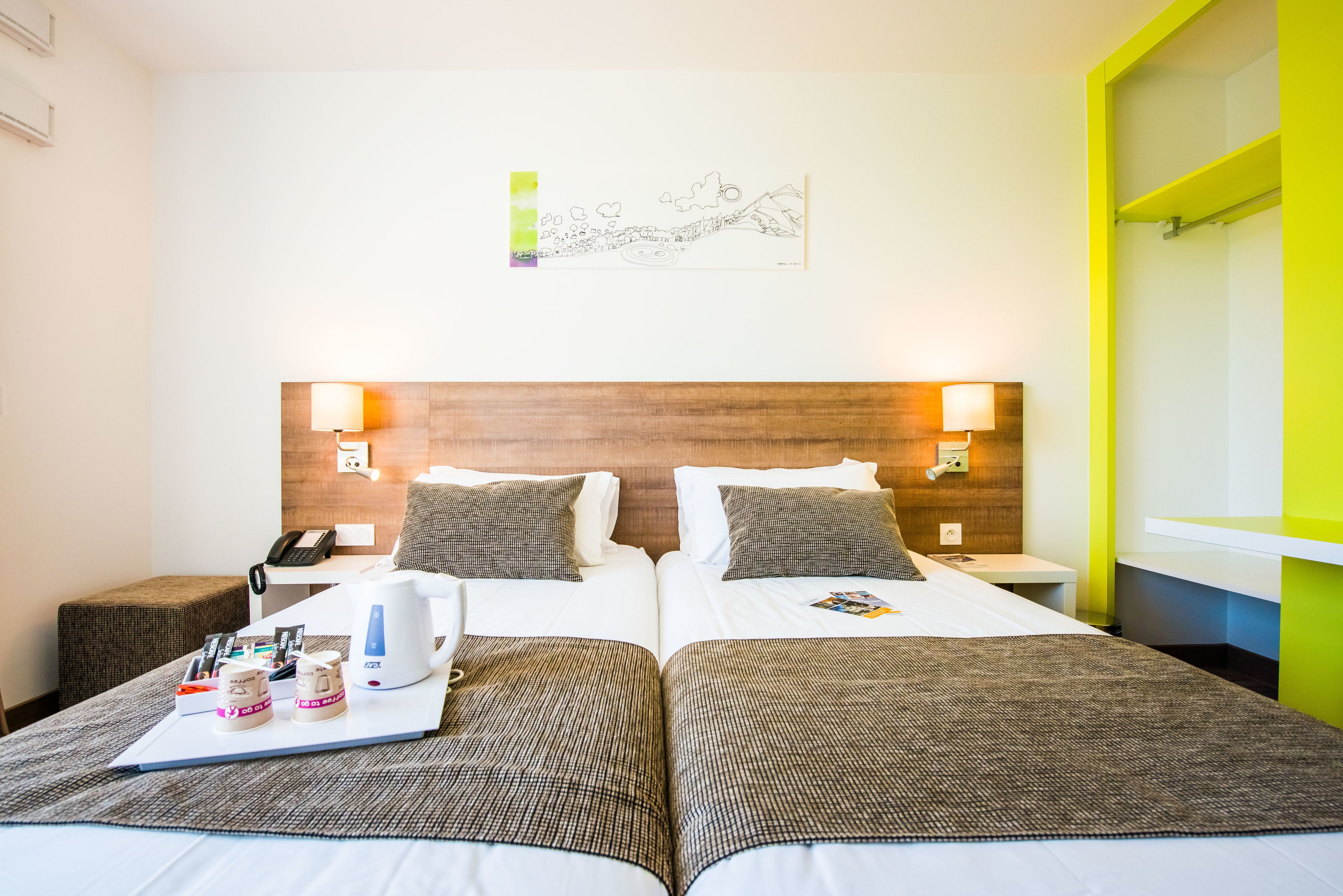 A hotel room with two beds, a bedside table with a lamp, a wall-mounted artwork, and a bright yellow accent wall.