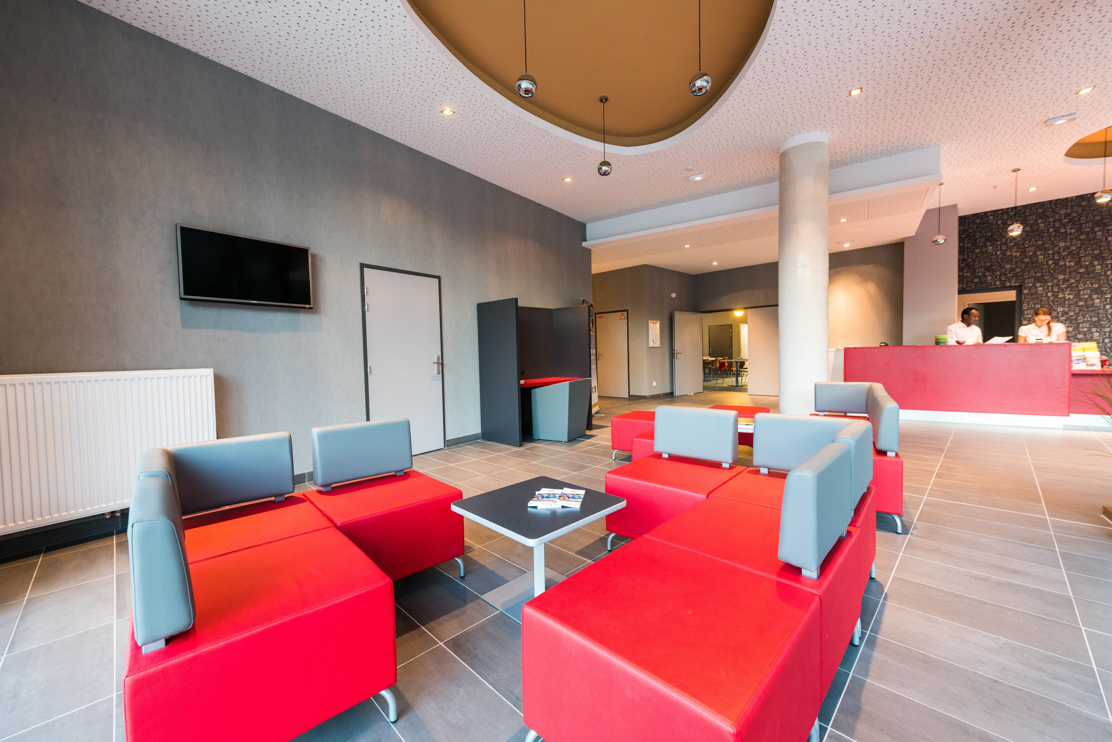 Een moderne lobby met rode en grijze zitplaatsen, een flatscreen-tv en een receptiebalie.