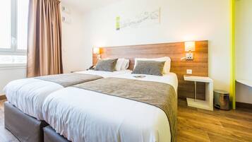 Camera d'albergo con un letto grande, due comodini, un tavolino, una lampada, un cestino e una finestra con tende.