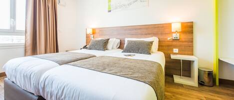 A hotel room with a large bed, two bedside tables, a nightstand, a lamp, a trash bin, and a window with curtains.