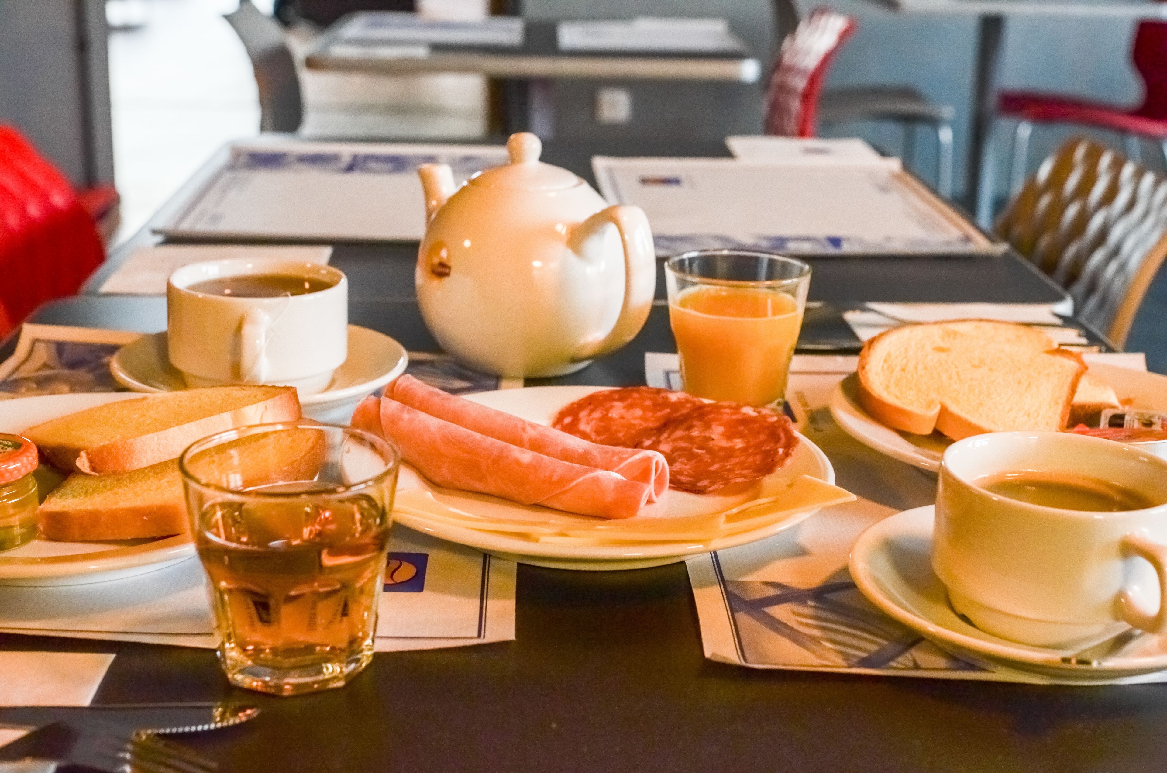 A breakfast setting with a teapot, cups, toast, sausages, and juice on a table.