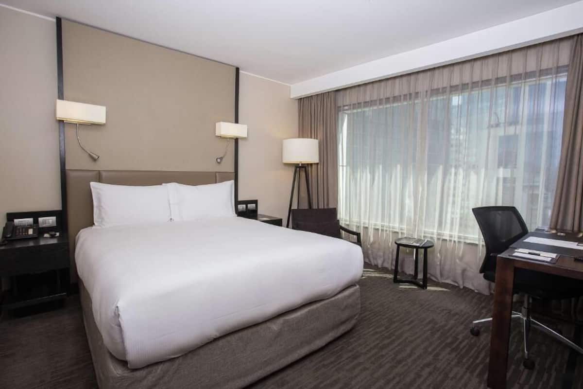 Ein Hotelzimmer mit einem großen Bett, zwei Nachttischen mit Lampen, einem Schreibtisch mit Stuhl und einem großen Fenster mit Vorhängen.