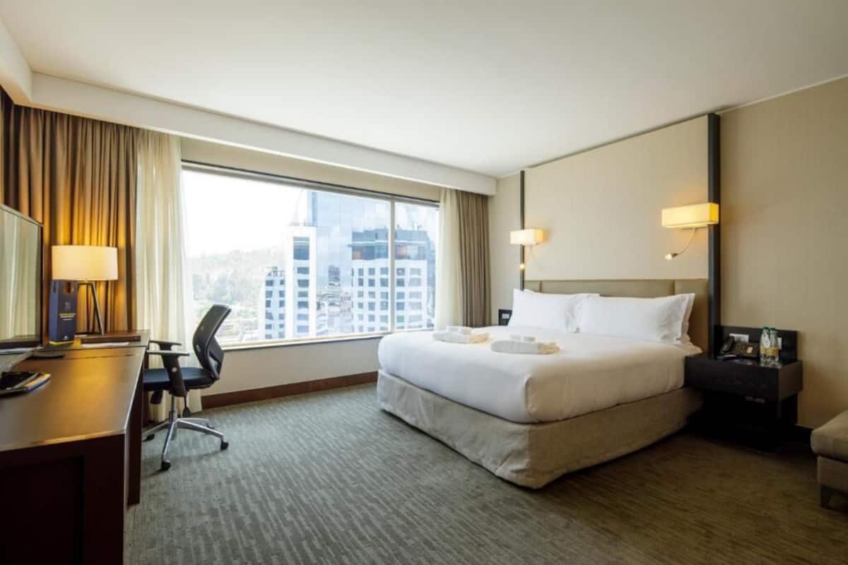 Ein Hotelzimmer mit einem großen Bett, einem Schreibtisch, einem Stuhl, einer Schreibtischlampe, einem Nachttisch und einem Fenster mit Blick auf die Stadt.