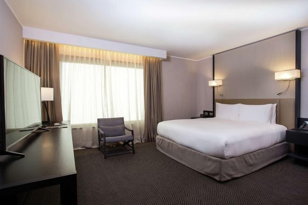Habitación de hotel con una cama grande, un escritorio con silla, un televisor y una ventana con cortinas.