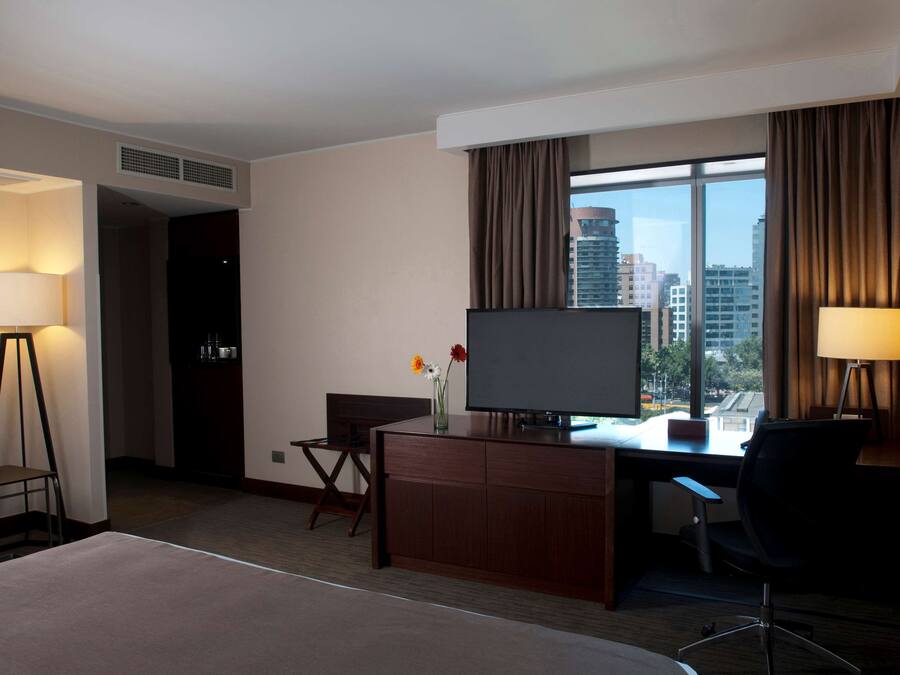 A hotel room with a bed, a desk with a TV, a chair, a lamp, and a window with curtains.