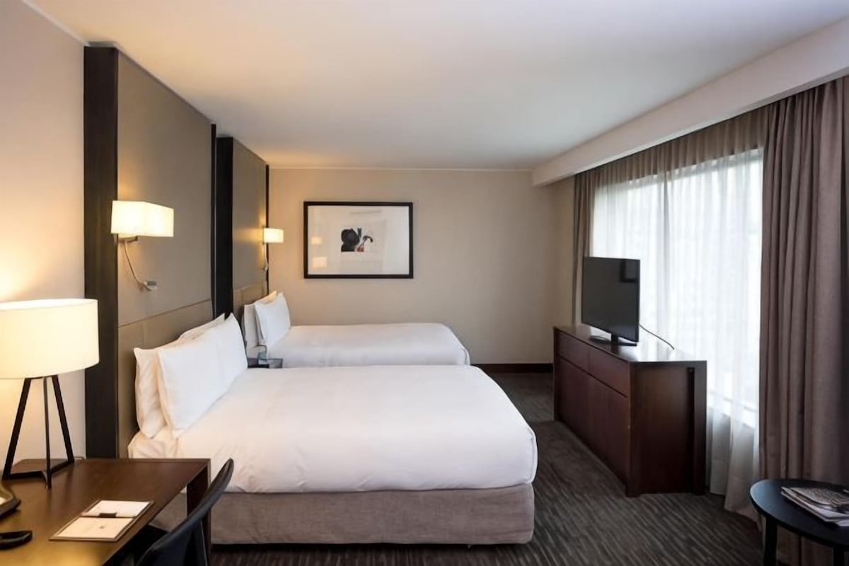 A hotel room with two beds, a desk, a chair, a TV, and a framed picture on the wall.