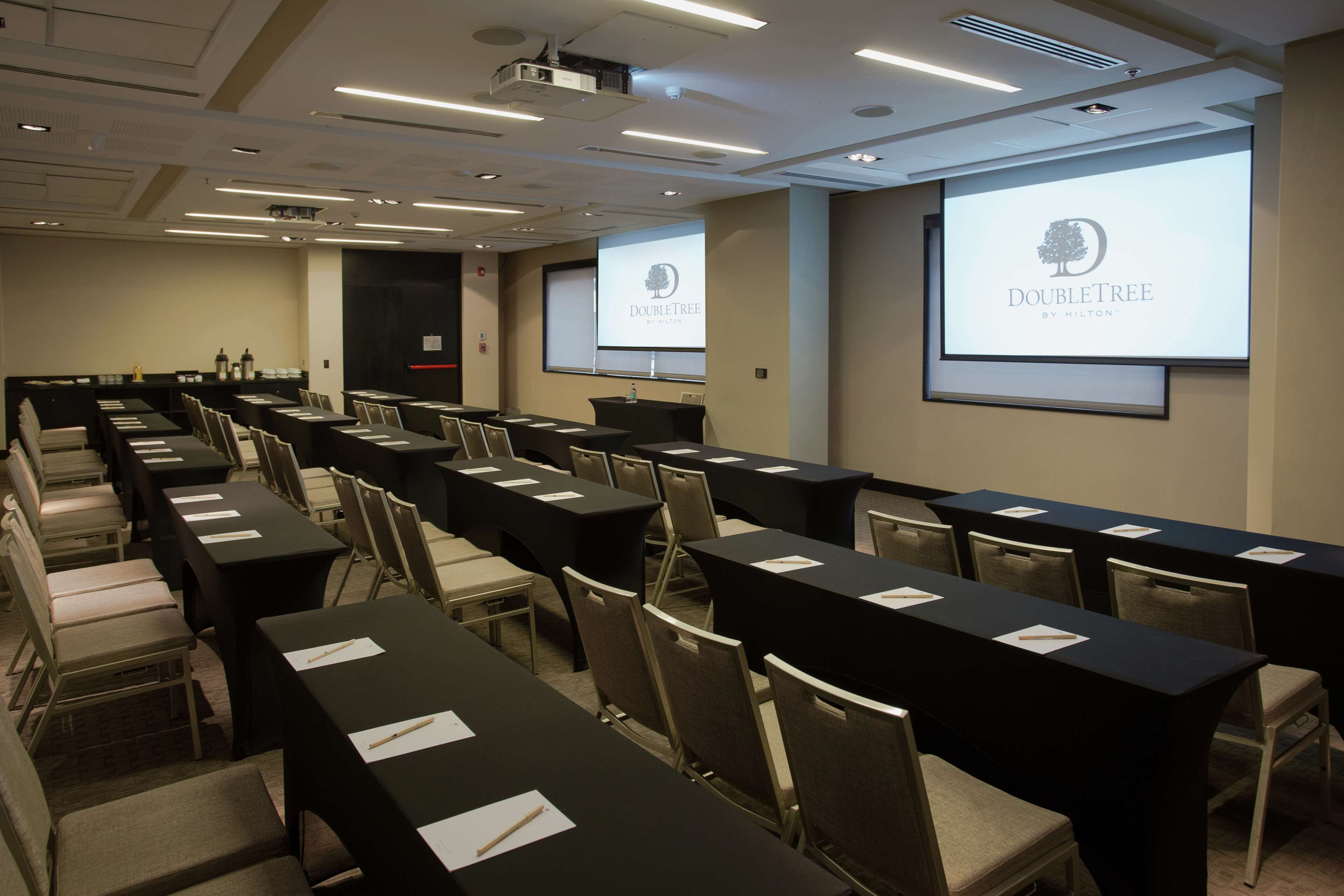 A conference room with rows of tables and chairs, a projector screen displaying 'DOUBLETREE by Hilton', and a presentation screen with the same logo.