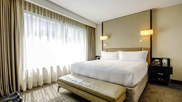Habitación de hotel con una cama grande, un reposapiés, una mesita de noche con lámpara y vistas al exterior a través de cortinas translúcidas.