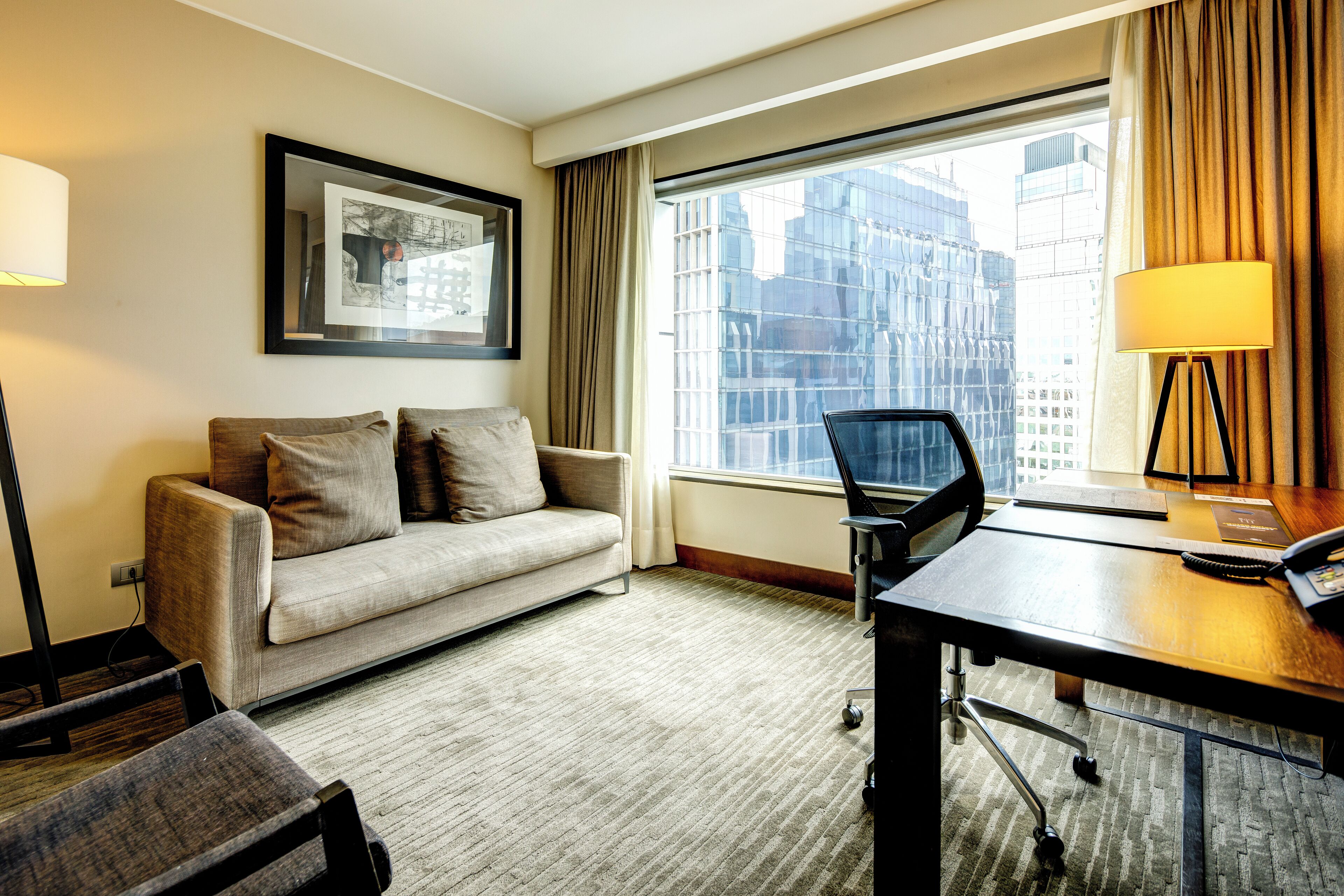 Ein Hotelzimmer mit Sofa, Schreibtisch, Stuhl, Lampe und einem großen Fenster mit Blick auf die Stadt.