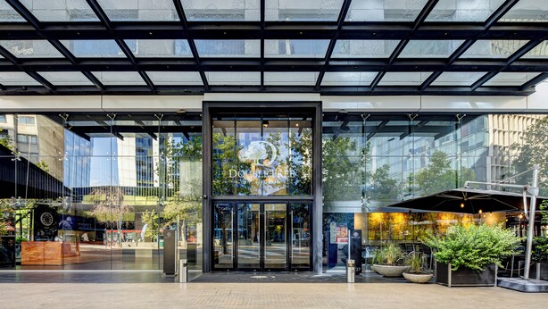 Una entrada de hotel moderna con fachada de vidrio, acceso central y zona de estar al aire libre.