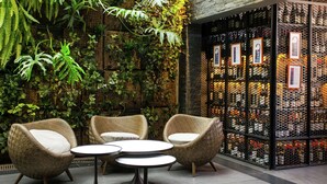 Cómodo espacio al aire libre con sillas de mimbre y mesas redondas, rodeado de abundante vegetación y un muro decorativo con fotografías.