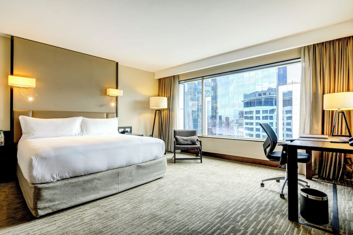 Ein Hotelzimmer mit einem großen Bett, einem Schreibtisch, einem Sessel und Blick auf eine Stadtlandschaft durch ein großes Fenster.