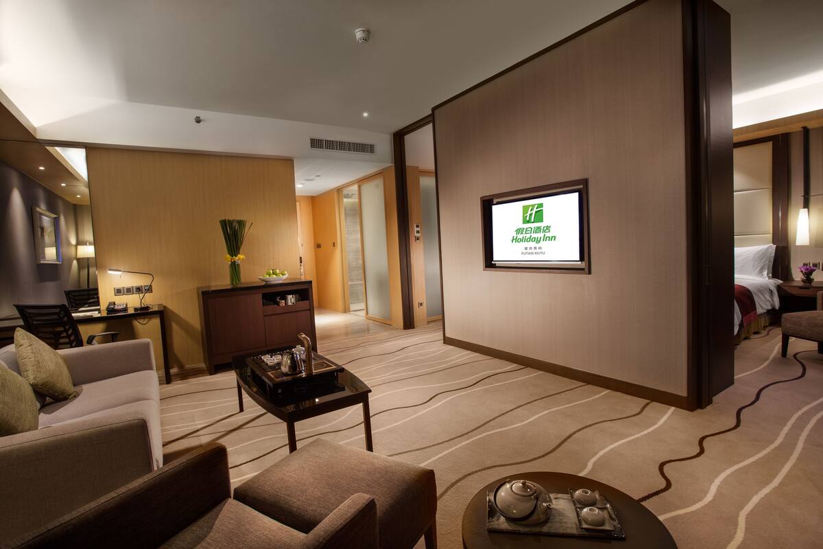 A hotel lobby with a sofa, armchairs, a coffee table, a TV screen, and a reception desk.
