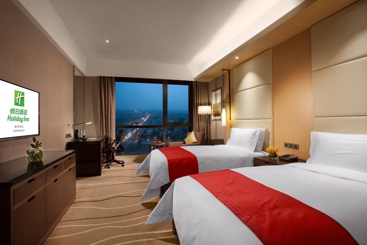 A hotel room with two beds, a desk, a TV, and a view of the city at dusk.