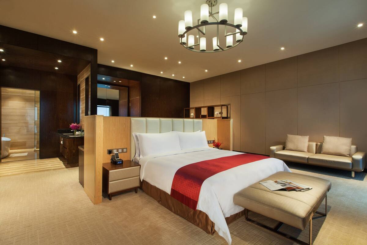 A modern hotel room with a large bed, a bedside table, a reading area, and a sofa.