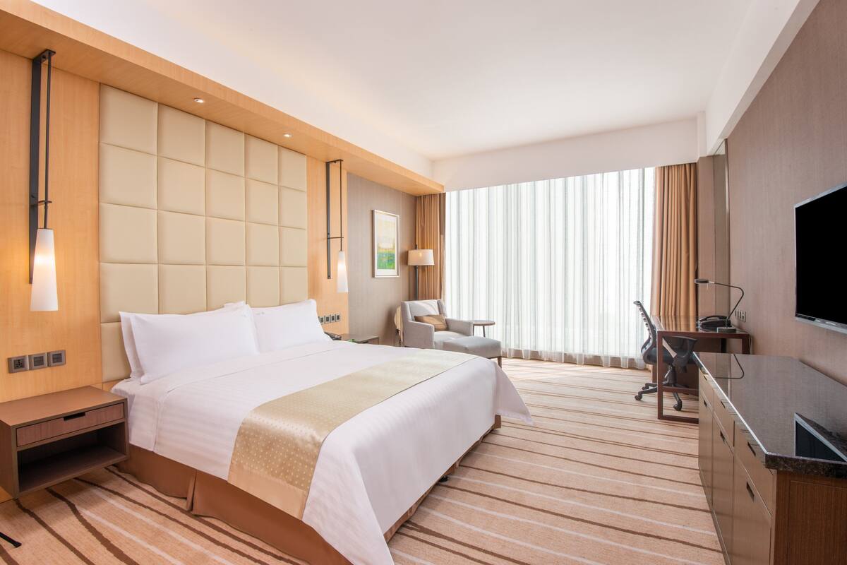 A hotel room with a large bed, a desk with a TV, a chair, and a window with curtains.
