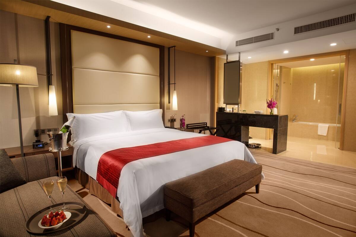 A hotel room with a large bed, a bedside table with a lamp, a small table with a plate of strawberries and two glasses of champagne, a bench, and a bathroom visible through an open door.