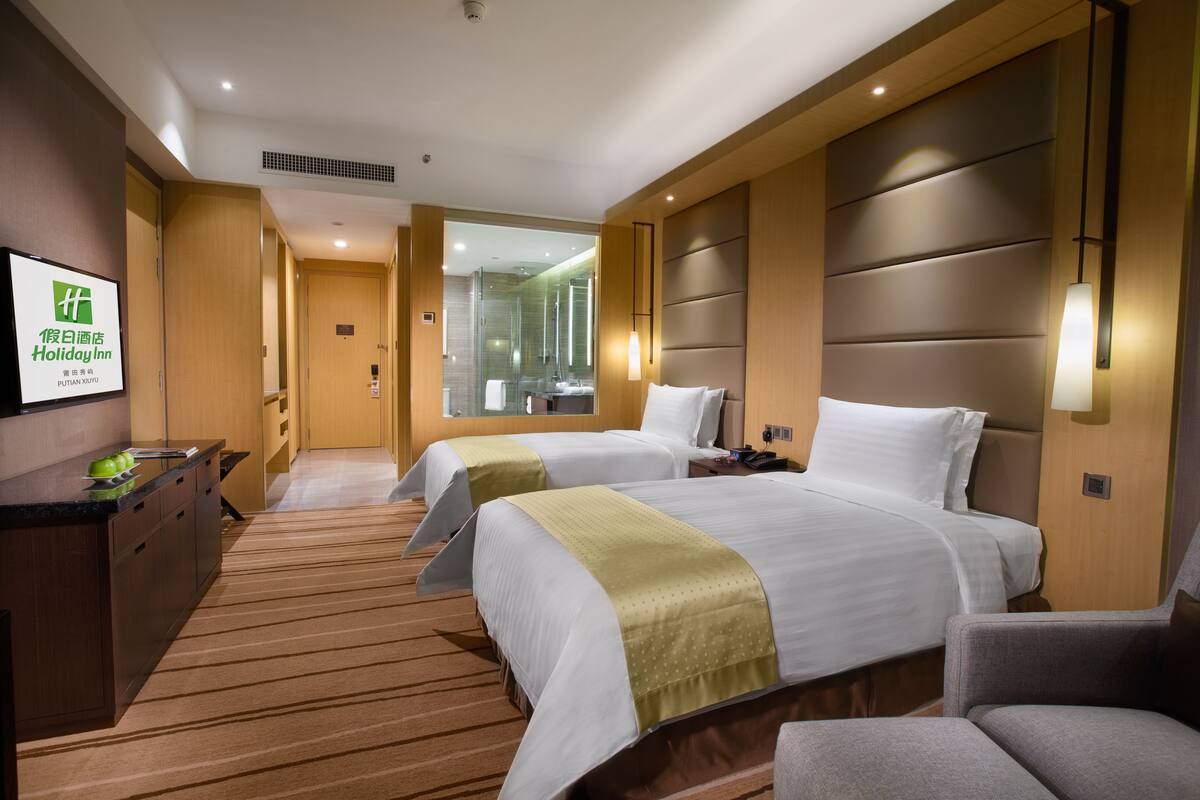 A hotel room with two beds, a desk, and a chair. There is a bathroom visible through a glass door.