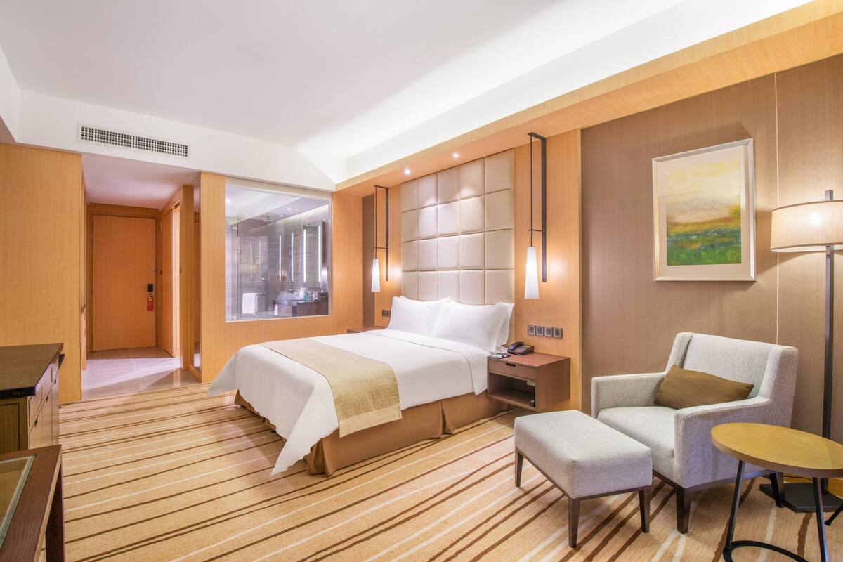 A hotel room with a large bed, a sitting area with a chair and ottoman, a small table, and a painting on the wall.