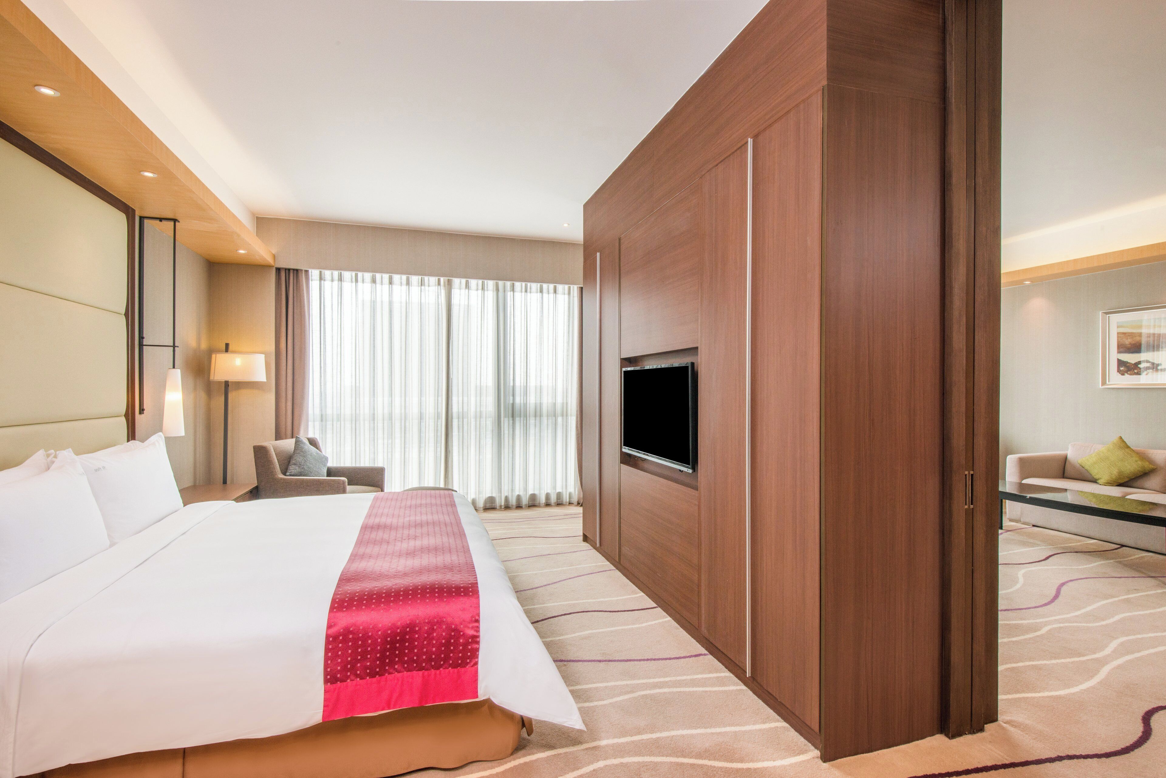 Habitación de hotel con una cama grande, un televisor de pantalla plana montado en la pared, una zona de estar con sofá y sillón, y un ventanal con cortinas traslúcidas.