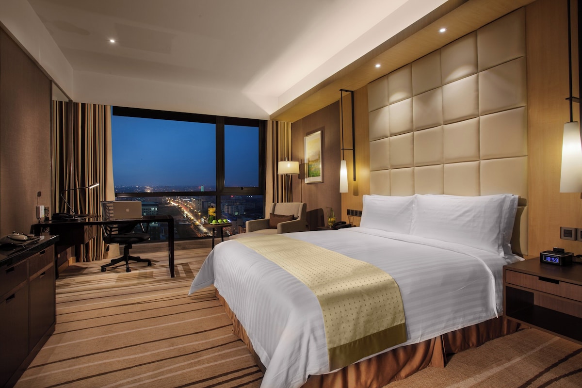 Habitación de hotel con una cama grande, un escritorio con una computadora portátil, vista de la ciudad de noche y un paisaje urbano visible a través de la ventana.