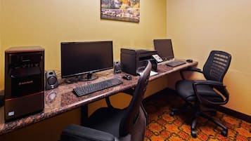 Una habitación de hotel con un escritorio equipado con una computadora de escritorio, un monitor, un teclado, un ratón e impresora. Hay dos sillas, una con respaldo y otra sin. Una cafetera está sobre el escritorio.