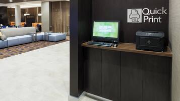 Un moderno vestíbulo de hotel con una mesa de recepción que incluye un monitor Lenovo, una impresora Dell y un área de descanso con un sofá y sillones.