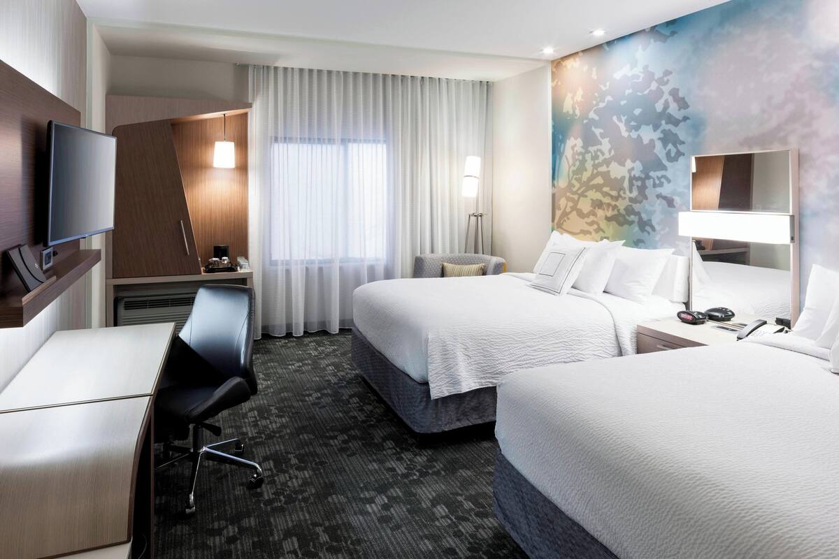A hotel room with two beds, a desk, a chair, a TV, and a large window with curtains.