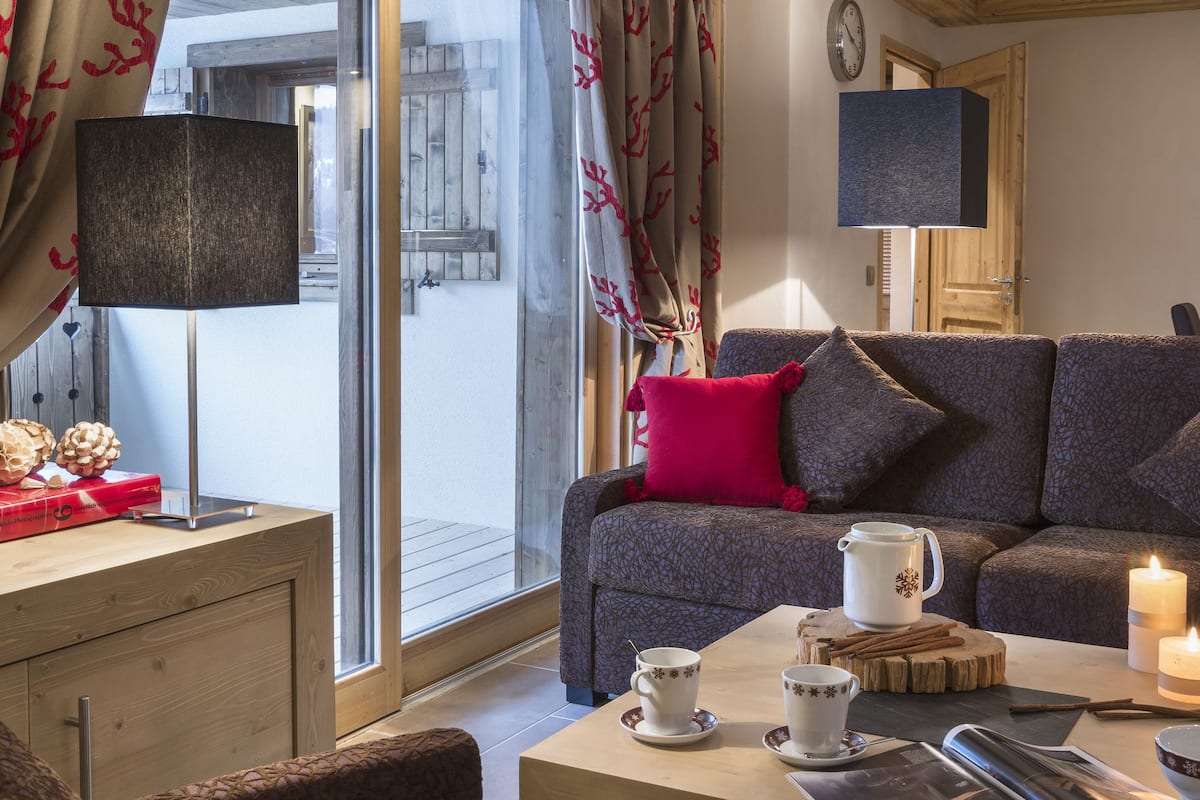 A cozy room with a sofa, a coffee table, and a TV. There is a window with curtains and a wooden ceiling.