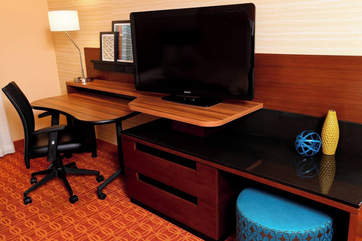 A hotel room with a wooden desk, a black office chair, a television, and a small blue ottoman.