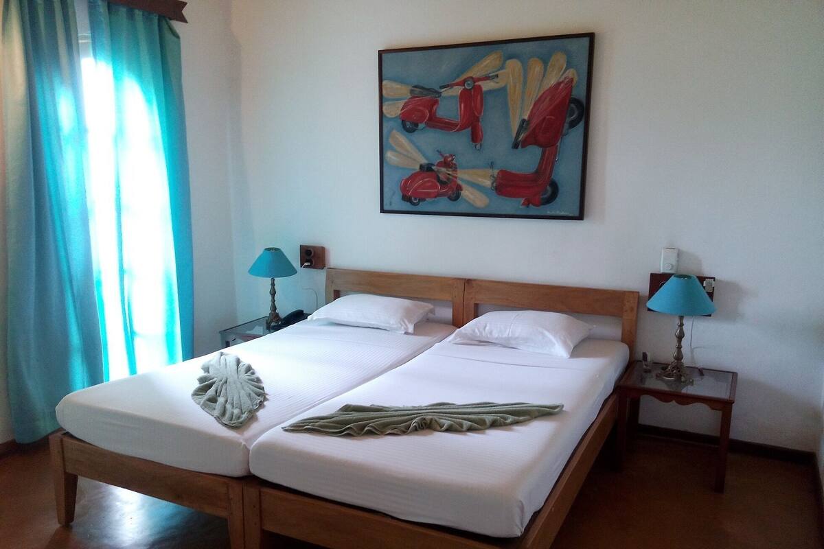 A double bed with white linens, two bedside tables with lamps, a painting on the wall, and a window with curtains.